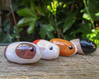 May include: Four painted rocks shaped like guinea pigs. The rocks are painted in different colors, including white, brown, orange, and black. The rocks have black eyes and are arranged in a row on a wooden surface.