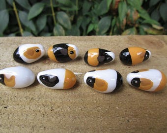 May include: Eight ceramic painted guinea pig figurines. The figurines are oval shaped and painted in black, white, and orange. The figurines are arranged in a row on a wooden surface.