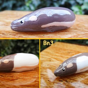 May include: Three hand-painted ceramic animal figurines, each depicting a small rodent with a white belly and brown head. The figurines are arranged in a row on a wooden surface. The first figurine is a dark gray color, the second is white with a brown head, and the third is white with a brown head.