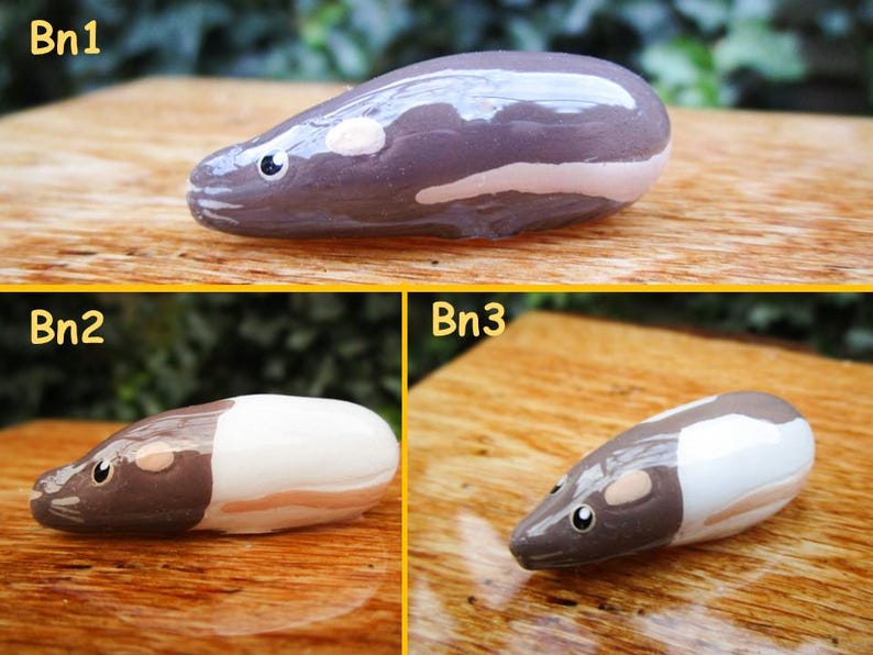 May include: Three hand-painted ceramic animal figurines, each depicting a small rodent with a white belly and brown head. The figurines are arranged in a row on a wooden surface. The first figurine is a dark gray color, the second is white with a brown head, and the third is white with a brown head.