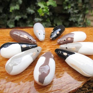 May include: A collection of nine ceramic mouse figurines, each painted in a different color combination of white, black, and brown. The mice are arranged in a circle on a wooden surface.