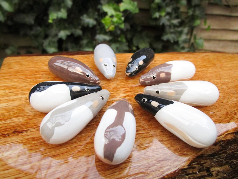 May include: A collection of nine ceramic mouse figurines, each painted in a different color combination of white, black, and brown. The mice are arranged in a circle on a wooden surface.