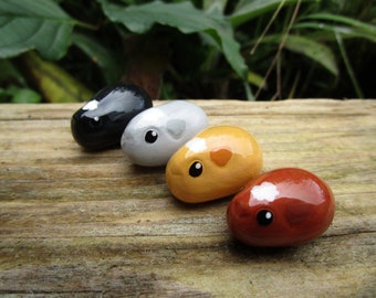 May include: Four painted stone figurines shaped like guinea pigs. The guinea pigs are painted in black, gray, orange, and red.