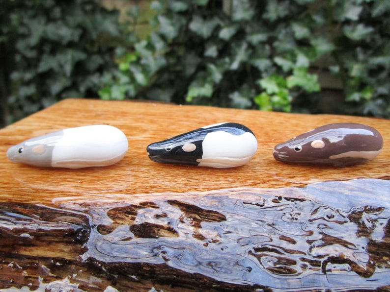 May include: Three ceramic animal figurines, each depicting a different type of rodent. The first is white with gray accents, the second is black and white, and the third is brown. The figurines are arranged in a row on a wooden surface.
