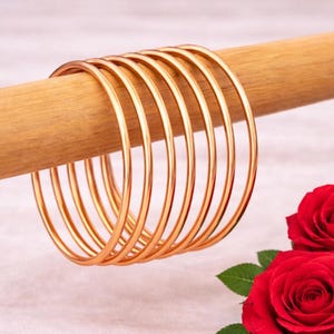 May include: A collection of gold-colored bangles displayed on a wooden dowel, with two vibrant red roses in the foreground. The bangles are thin and circular, reflecting light. The roses have multiple petals and green leaves.