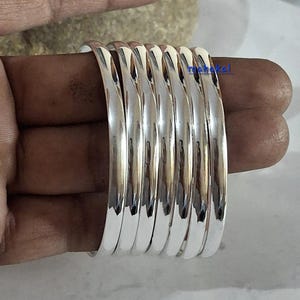 Può includere: Una pila di sette braccialetti argentati lucenti. I braccialetti hanno una superficie liscia e riflettente, creando un aspetto metallico brillante. I braccialetti circolari sono impilati. La parola "mahakal" è visibile sui braccialetti.