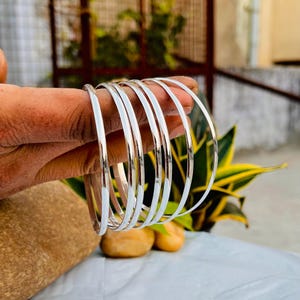 Op de afbeelding: Een set van tien zilveren armbanden, gestapeld en gerangschikt in een cirkelvorm. De armbanden zijn glad en glanzend, met een licht afgeronde rand.