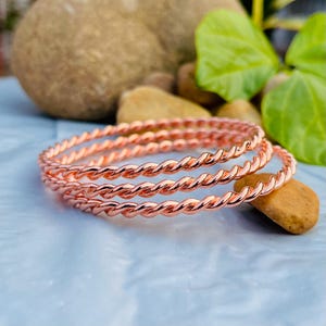 May include: Three rose gold colored twisted bangle bracelets. The bracelets are stacked on top of each other.