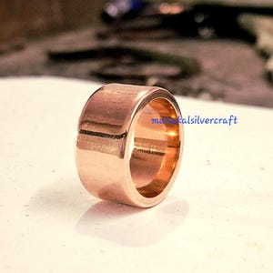Puede incluir: Un anillo pulido de oro rosa con una banda ancha. El anillo tiene una superficie lisa y reflectante y un diseño simple y minimalista. El texto "mahikalsilvercraft" es visible en el anillo.