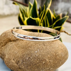 May include: A simple silver bangle bracelet with a polished finish. The bracelet is resting on a brown rock.