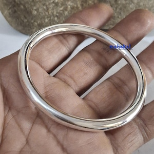 May include: A smooth, polished silver-toned bangle bracelet with a rounded, thick profile. The surface of the metal is reflective and plain. The word mahakal is printed in small blue letters on the upper right side of the circular metal band.