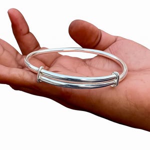 Könnte beinhalten: Ein glänzendes, verstellbares Armreif-Armband aus Silber. Das Armband hat ein schlichtes Design mit glatter, polierter Oberfläche. Die verstellbare Funktion ermöglicht eine bequeme Passform. Ein klassisches Schmuckstück.