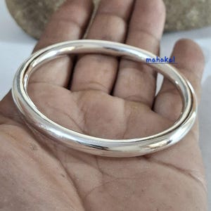 Könnte beinhalten: Ein glänzendes, silbernes Armreif-Armband mit einem glatten, abgerundeten Design. Das Armband ist kreisförmig und scheint aus Metall zu sein. Das Wort "mahakal" ist auf dem Armband aufgedruckt.