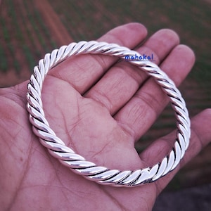 Puede incluir: Un brazalete plateado con un diseño de cuerda trenzada. El brazalete circular parece ser de metal. La palabra "mahakal" es visible en la imagen. El brazalete se sostiene en una mano.