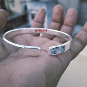 Könnte beinhalten: Ein silberfarbenes, offenes Armreif-Armband mit dem Wort "mahakal" in Rot. Das Armband wird in einer Hand gehalten, wodurch seine polierte Oberfläche hervorgehoben wird. Der Schmuck ist schlicht und elegant.