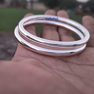 May include: Two silver bangles, stacked, with a smooth, reflective surface. The word "mahakal" is printed on the top bangle. The bangles are held in a hand, with a blurred green background.