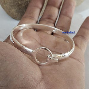 Könnte beinhalten: Ein silberfarbenes Armreif-Armband mit einem kreisförmigen Verschluss. Das Armband ist glatt und glänzend, mit einem schlichten, eleganten Design. Das Wort "mahakal" ist auf dem Armband aufgedruckt.