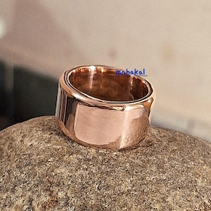 May include: A rose gold ring with a wide band. The ring has a smooth, polished surface and a simple, modern design. The ring is displayed on a textured, gray stone.