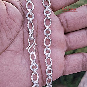 Puede incluir: Collar plateado con diseño de eslabones ovalados texturizados. El collar tiene un cierre de gancho. La palabra "Galaxy" es visible en la esquina superior derecha. Se muestra sobre un fondo neutro.