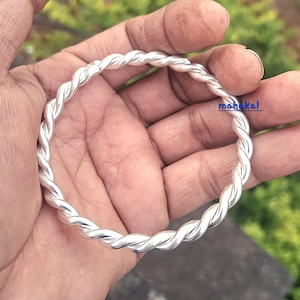 May include: A silver bangle with a twisted rope design. The bracelet is held in a hand, with the word "mahakal" visible on the bangle. The background is a blurred green.
