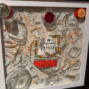 May include: A framed artwork featuring a broken glass bottle with a label that reads "Tanqueray Flor de Sevilla Distilled Gin". The bottle is surrounded by shards of glass and orange glitter.