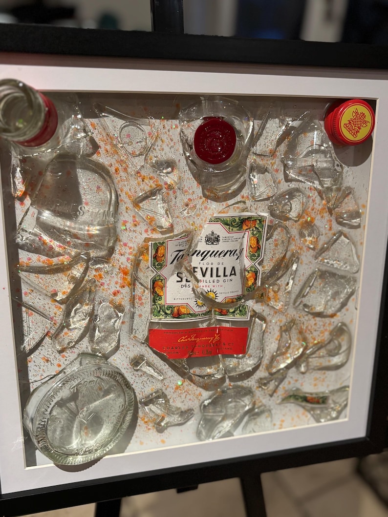 May include: A framed artwork featuring a broken glass bottle with a label that reads "Tanqueray Flor de Sevilla Distilled Gin". The bottle is surrounded by shards of glass and orange glitter.