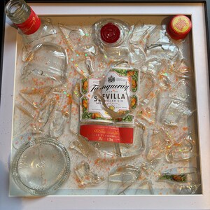 May include: A framed artwork featuring a Tanqueray Sevilla Orange Flavored Gin bottle label surrounded by broken glass pieces and glitter. The artwork is set against a white background.