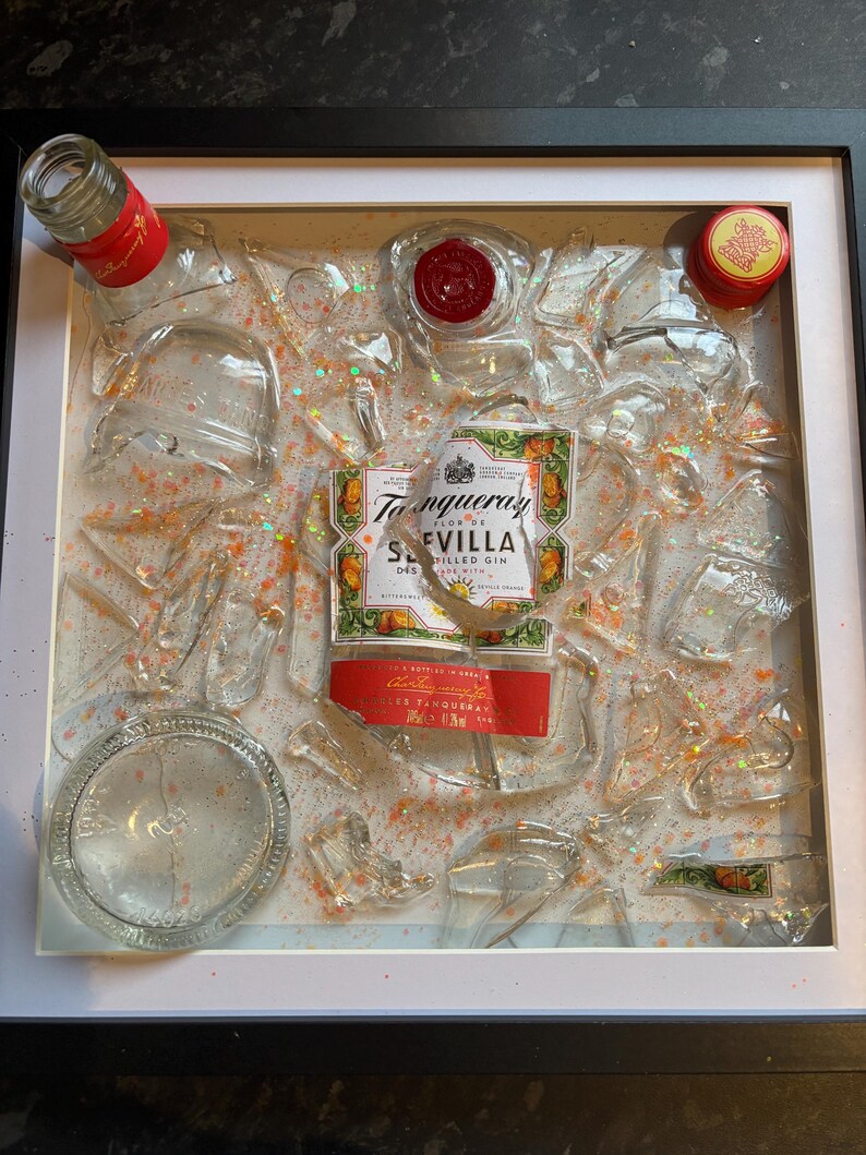 May include: A framed artwork featuring a Tanqueray Sevilla Orange Flavored Gin bottle label surrounded by broken glass pieces and glitter. The artwork is set against a white background.