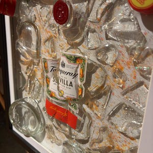 May include: A framed artwork featuring a broken glass bottle of Tanqueray Sevilla Dry Gin. The bottle is surrounded by pieces of broken glass and a white background with orange and yellow accents.