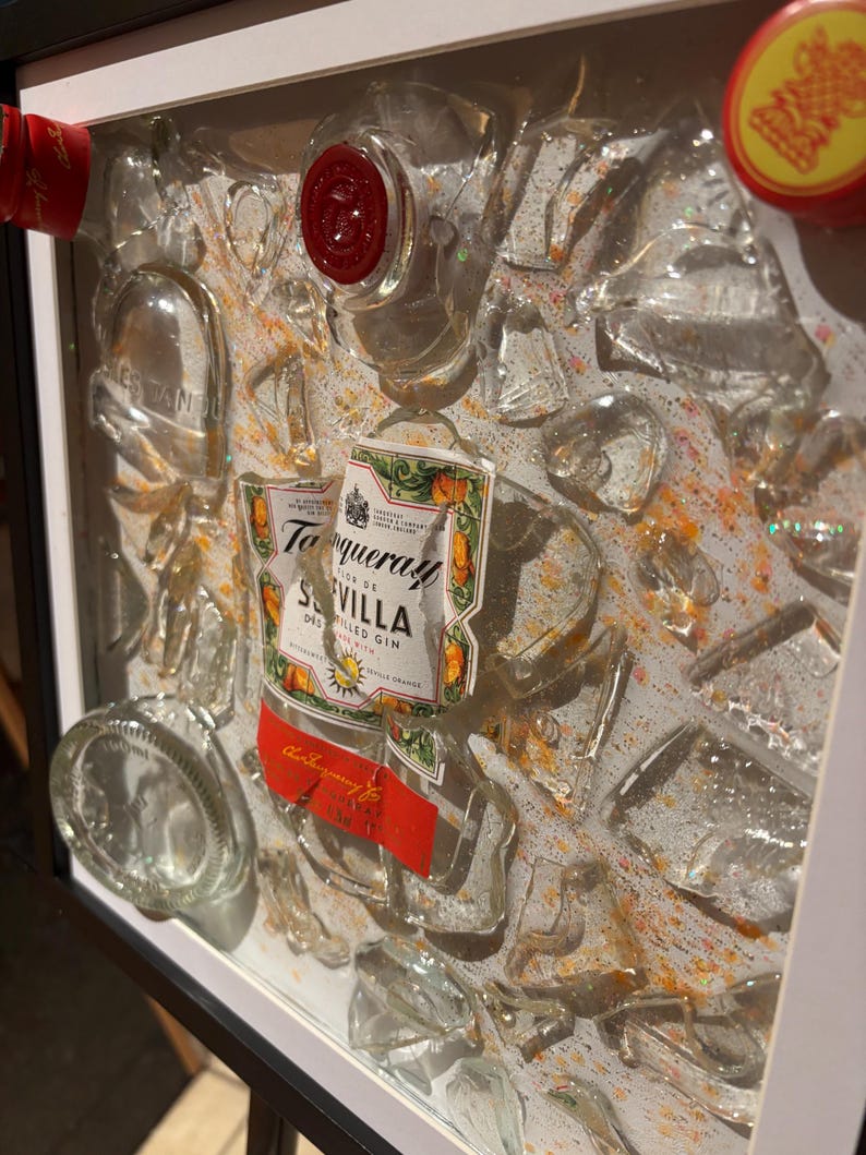May include: A framed artwork featuring a broken glass bottle of Tanqueray Sevilla Dry Gin. The bottle is surrounded by pieces of broken glass and a white background with orange and yellow accents.
