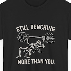 Puede incluir: Camiseta negra con un gráfico blanco de un esqueleto levantando pesas. El texto encima del esqueleto dice "STILL BENCHING" y debajo dice "MORE THAN YOU."