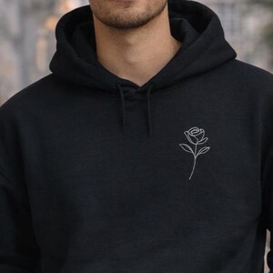 Könnte beinhalten: Schwarzer Kapuzenpullover mit weißer Rosenkontur auf der Brust. Der Kapuzenpullover hat eine Kapuze mit Kordelzug und eine Kängurutasche vorne. Das Bild zeigt eine Person, die den Kapuzenpullover trägt. Der Kapuzenpullover ist aus weichem Material.