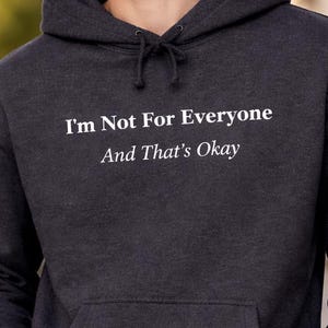 Puede incluir: Sudadera con capucha gris oscuro con el texto blanco "I'm Not For Everyone And That's Okay". La sudadera tiene una capucha con cordón y un bolsillo delantero. La persona lleva vaqueros negros.