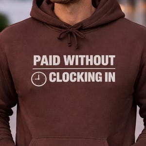 May include: A brown hoodie with the text "PAID WITHOUT CLOCKING IN" in white. The hoodie has a front pocket and a drawstring hood. The person in the image is wearing blue jeans and a watch.