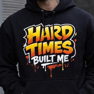May include: Black hooded sweatshirt with the text "HARD TIMES BUILT ME" in a bold, stylized font. The text is primarily yellow and orange with white and red accents. The sweatshirt has a front pocket and a drawstring hood.