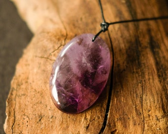 May include: A purple amethyst gemstone pendant on a black cord necklace. The pendant is oval shaped and has a smooth, polished surface.
