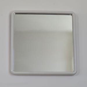Op de afbeelding: Vierkante spiegel met een afgeronde, lichtgrijze lijst. De spiegel reflecteert de omgeving. De lijst is effen van kleur en heeft een gladde afwerking. De spiegel is aan een witte muur bevestigd.
