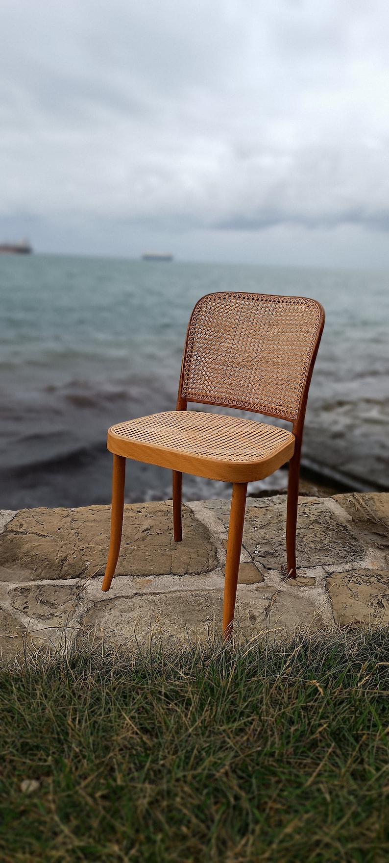 May include: A wooden chair with a woven cane seat and back. The chair is sitting on a stone patio with green grass in the foreground.