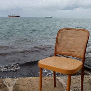May include: A wooden chair with a woven cane seat and back sits on a stone patio overlooking the ocean. Two ships are visible in the distance.