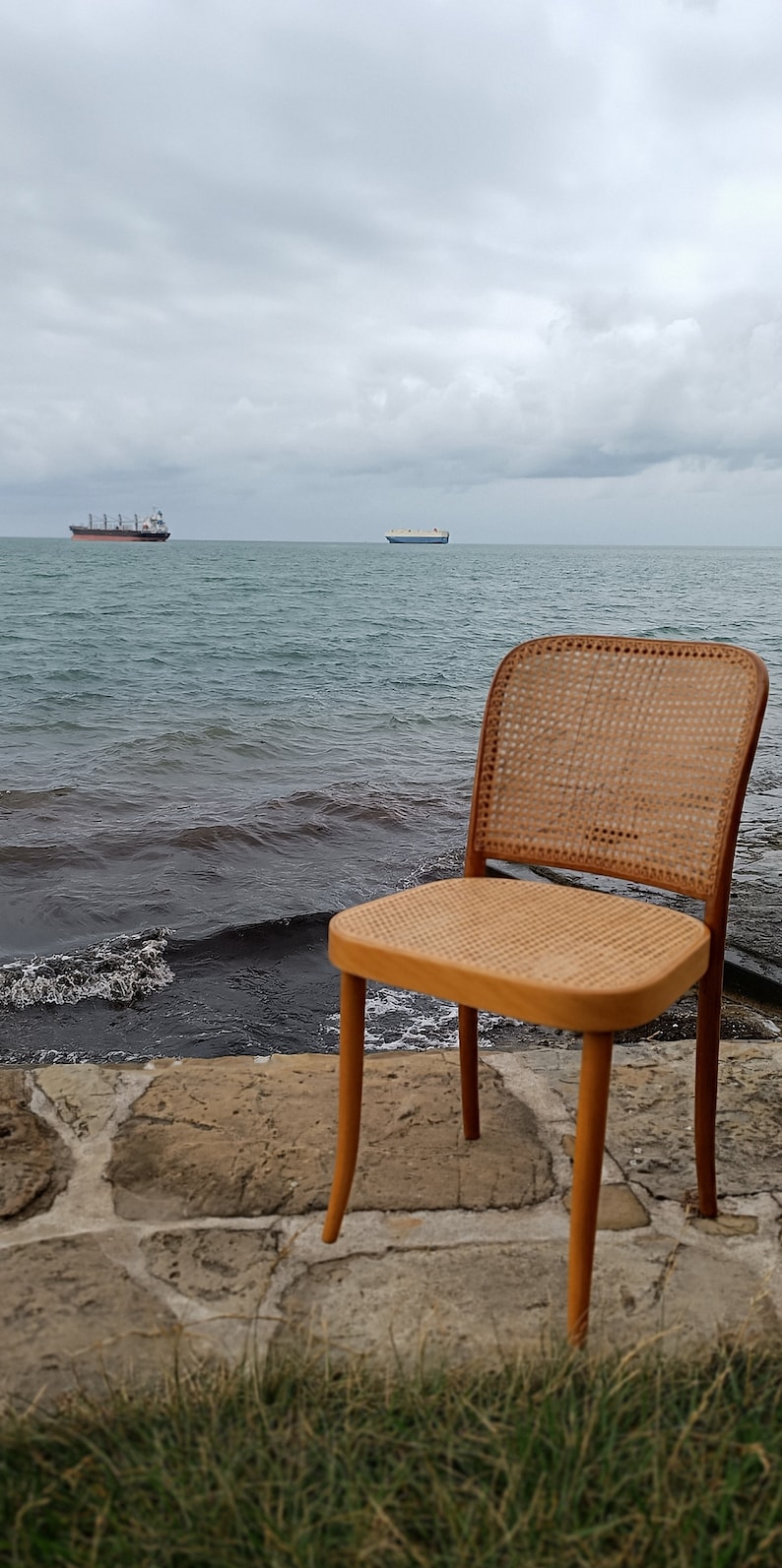 May include: A wooden chair with a woven cane seat and back sits on a stone patio overlooking the ocean. Two ships are visible in the distance.