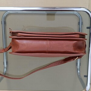 Pode incluir: Uma bolsa transversal de couro marrom com uma al&ccedil;a longa e ajust&aacute;vel. A bolsa possui um fecho de z&iacute;per superior e formato retangular. O couro parece liso e tem um tom marrom-avermelhado quente.