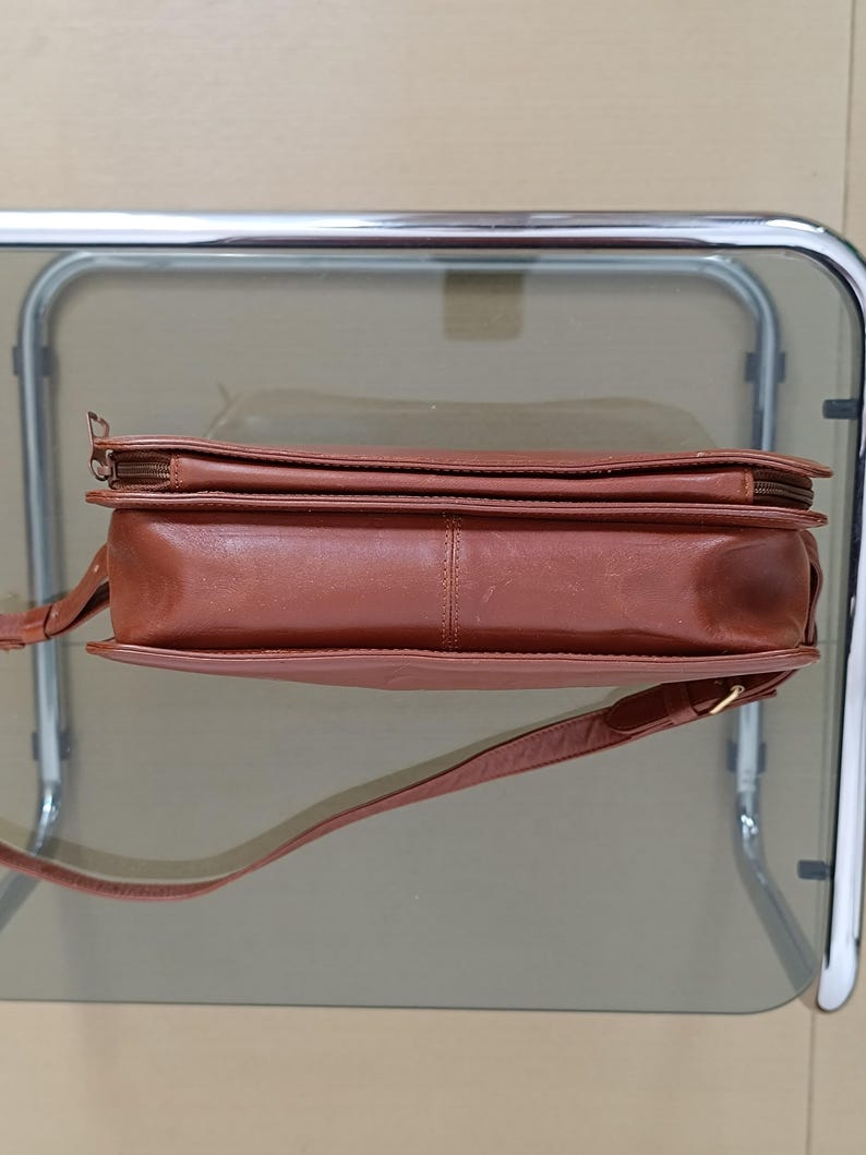 Pode incluir: Uma bolsa transversal de couro marrom com uma al&ccedil;a longa e ajust&aacute;vel. A bolsa possui um fecho de z&iacute;per superior e formato retangular. O couro parece liso e tem um tom marrom-avermelhado quente.