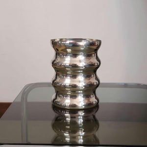 May include: A silver metal vase with a wavy, rippled design. The vase is sitting on a glass table.