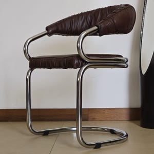 May include: A brown leather chair with a chrome frame. The chair has a curved back and a padded seat. The chair is on a tiled floor.