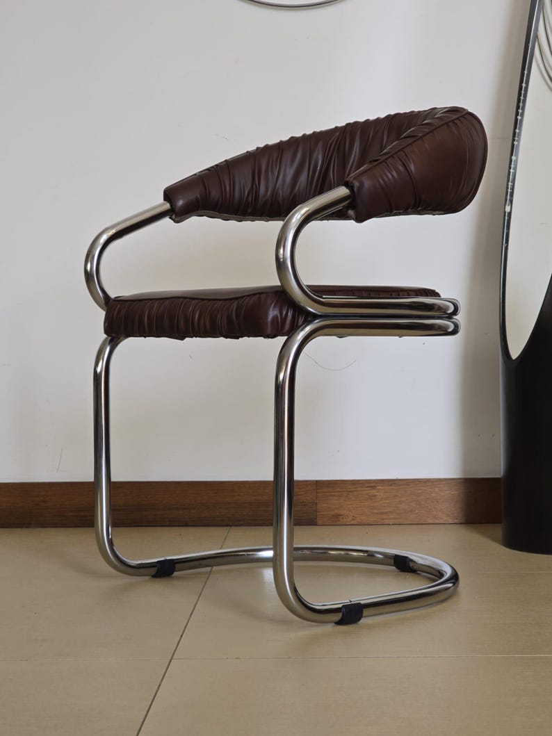 May include: A brown leather chair with a chrome frame. The chair has a curved back and a padded seat. The chair is on a tiled floor.
