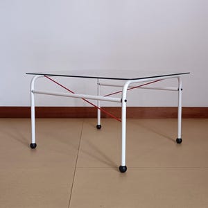 Könnte beinhalten: Couchtisch mit weißer Glasplatte, weißem Metallgestell und roten Gummibändern. Der Tisch hat vier schwarze Rollen.