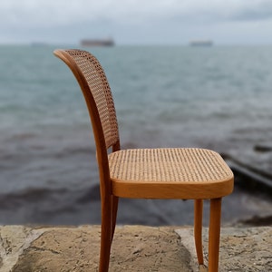 May include: A wooden chair with a woven cane seat and back. The chair is sitting on a stone patio with grass in the foreground. The background is a blurry image of a body of water.