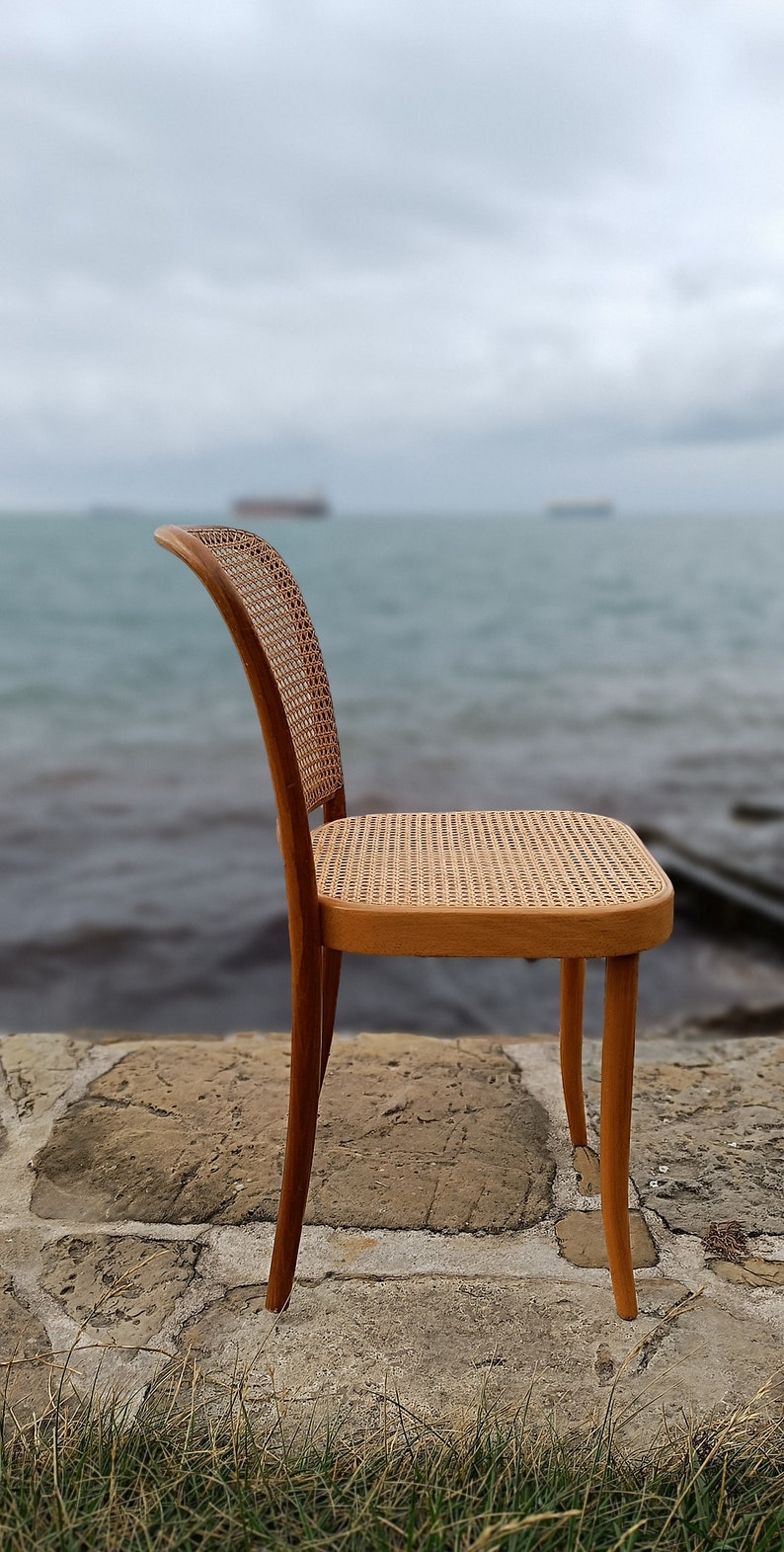 May include: A wooden chair with a woven cane seat and back. The chair is sitting on a stone patio with grass in the foreground. The background is a blurry image of a body of water.