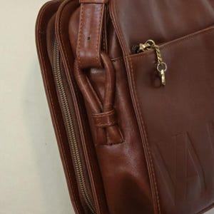 Pode incluir: Bolsa de ombro de couro castanho com fecho de aba e compartimento com z&iacute;per. A bolsa apresenta uma longa al&ccedil;a ajust&aacute;vel e um z&iacute;per dourado. A palavra "VAL" est&aacute; em relevo na frente da bolsa.