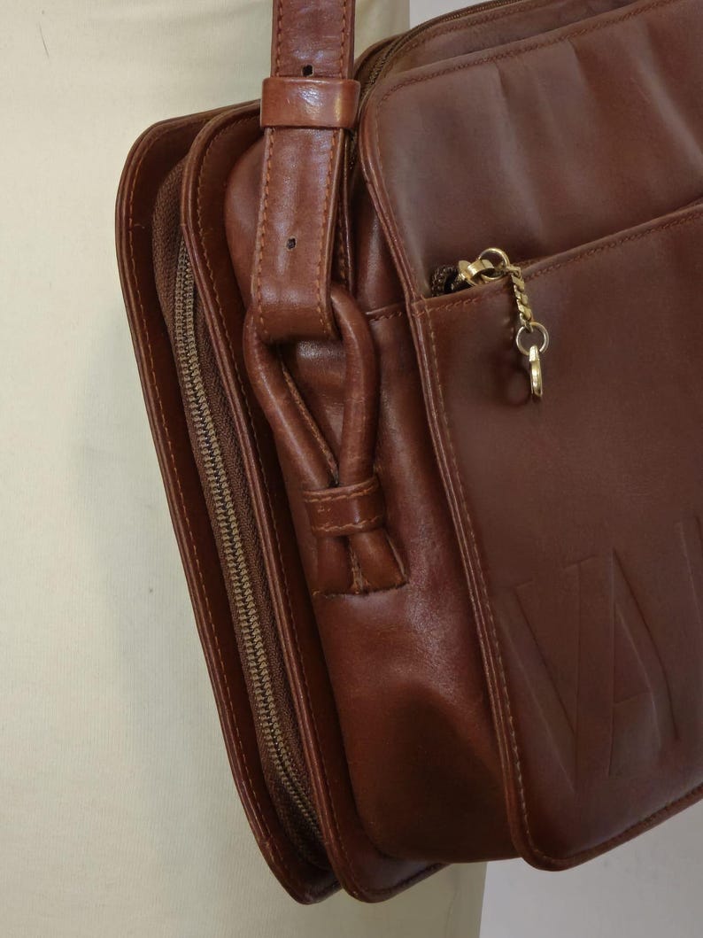 Pode incluir: Bolsa de ombro de couro castanho com fecho de aba e compartimento com z&iacute;per. A bolsa apresenta uma longa al&ccedil;a ajust&aacute;vel e um z&iacute;per dourado. A palavra "VAL" est&aacute; em relevo na frente da bolsa.